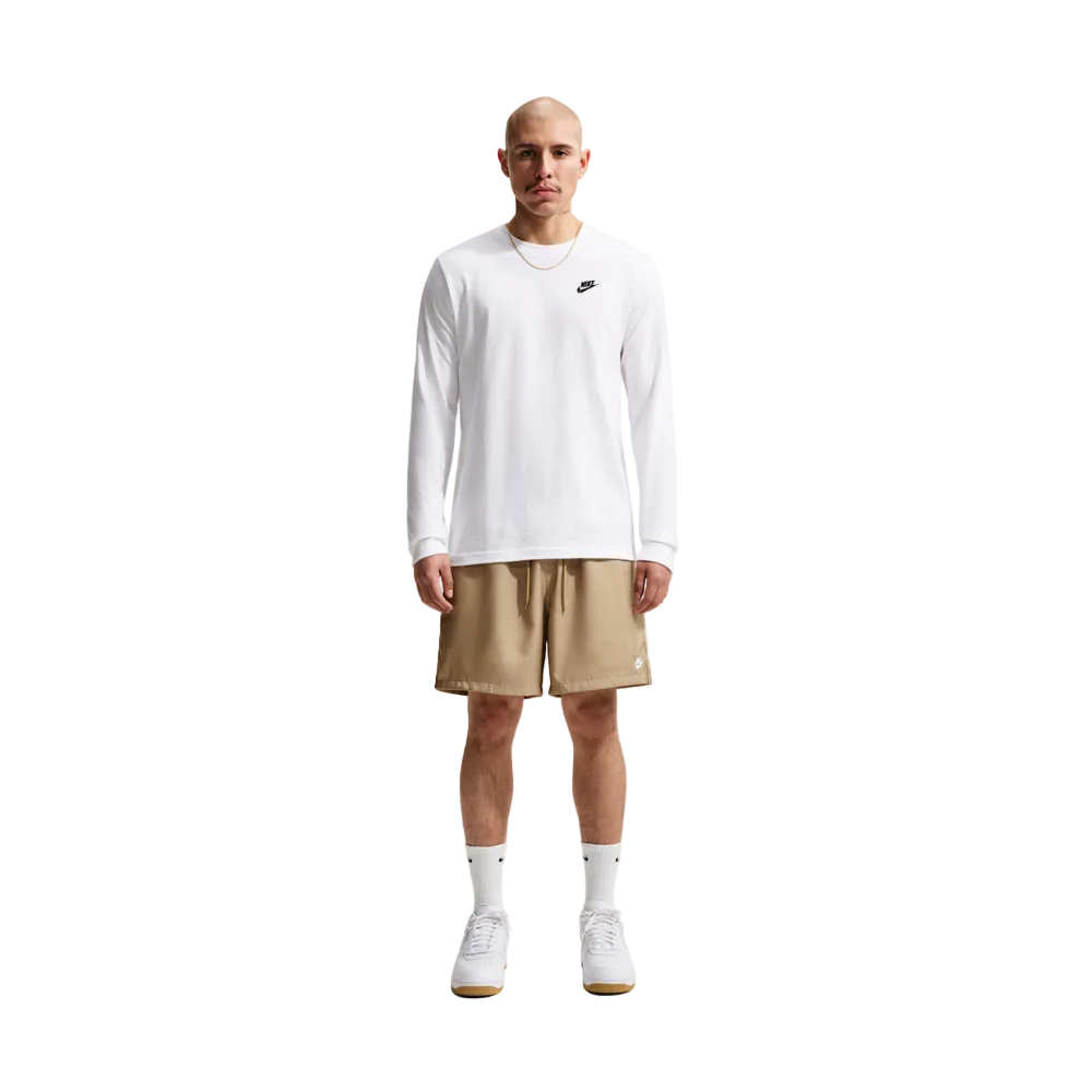 A man stands against a light gray studio background wearing a white long-sleeve Nike shirt, tan athletic shorts, white socks, and white sneakers.