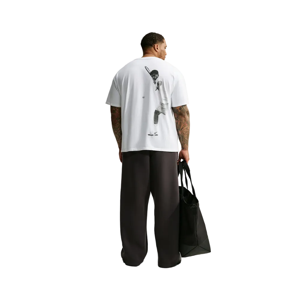 A man stands with his back to the camera wearing a white oversized T-shirt with a large black graphic print on the back, loose dark pants, and holding a black tote bag against a plain light gray studio background.