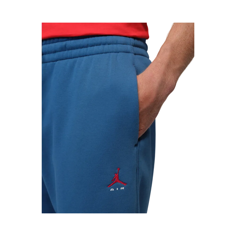 Close-up of a person wearing blue sweatpants with a red embroidered Jordan logo near the lower left leg, with a red shirt partially visible at the top against a light gray background.