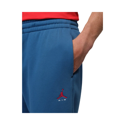 Close-up of a person wearing blue sweatpants with a red embroidered Jordan logo near the lower left leg, with a red shirt partially visible at the top against a light gray background.