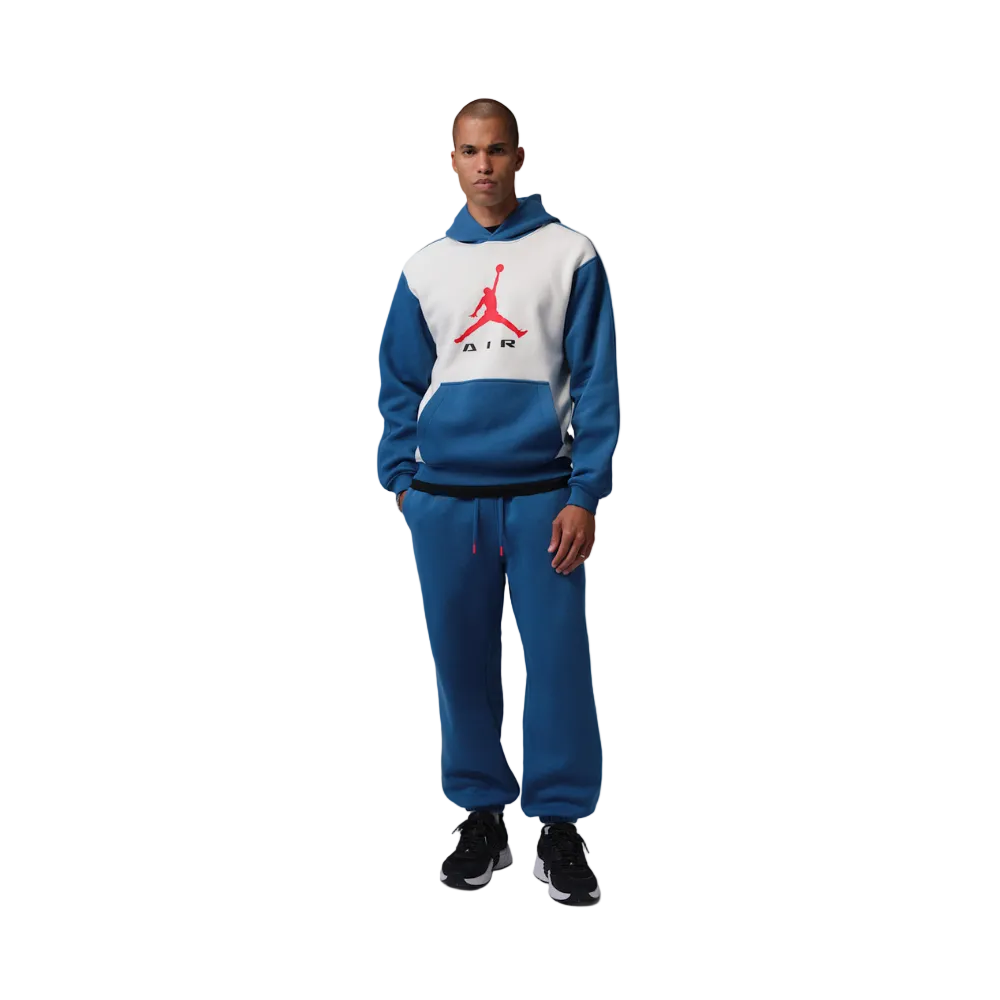A male model stands against a light gray studio background wearing a blue and white color-blocked hoodie with a red Jumpman logo and matching blue sweatpants.