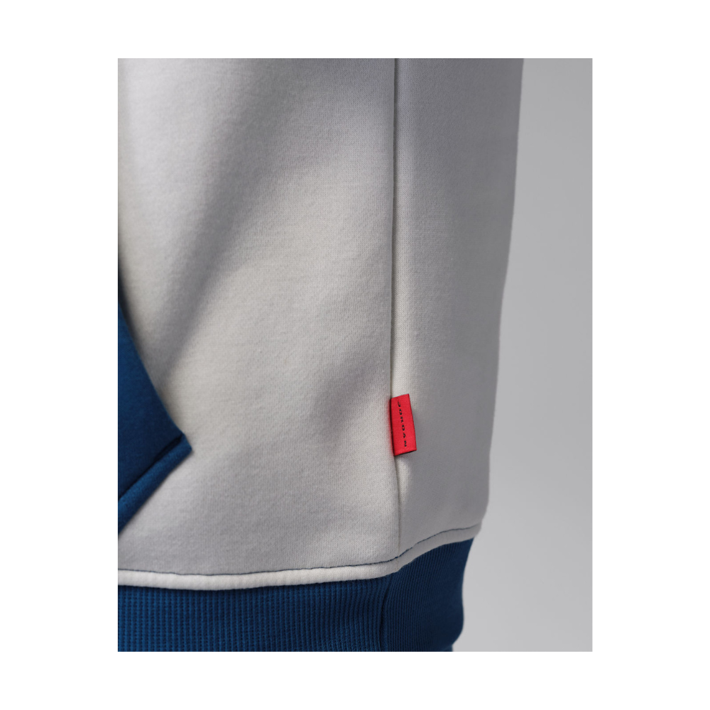 Close-up of a light gray sweatshirt with a small red brand tab on the front, paired with a dark blue ribbed hem, shown against a neutral gray background.