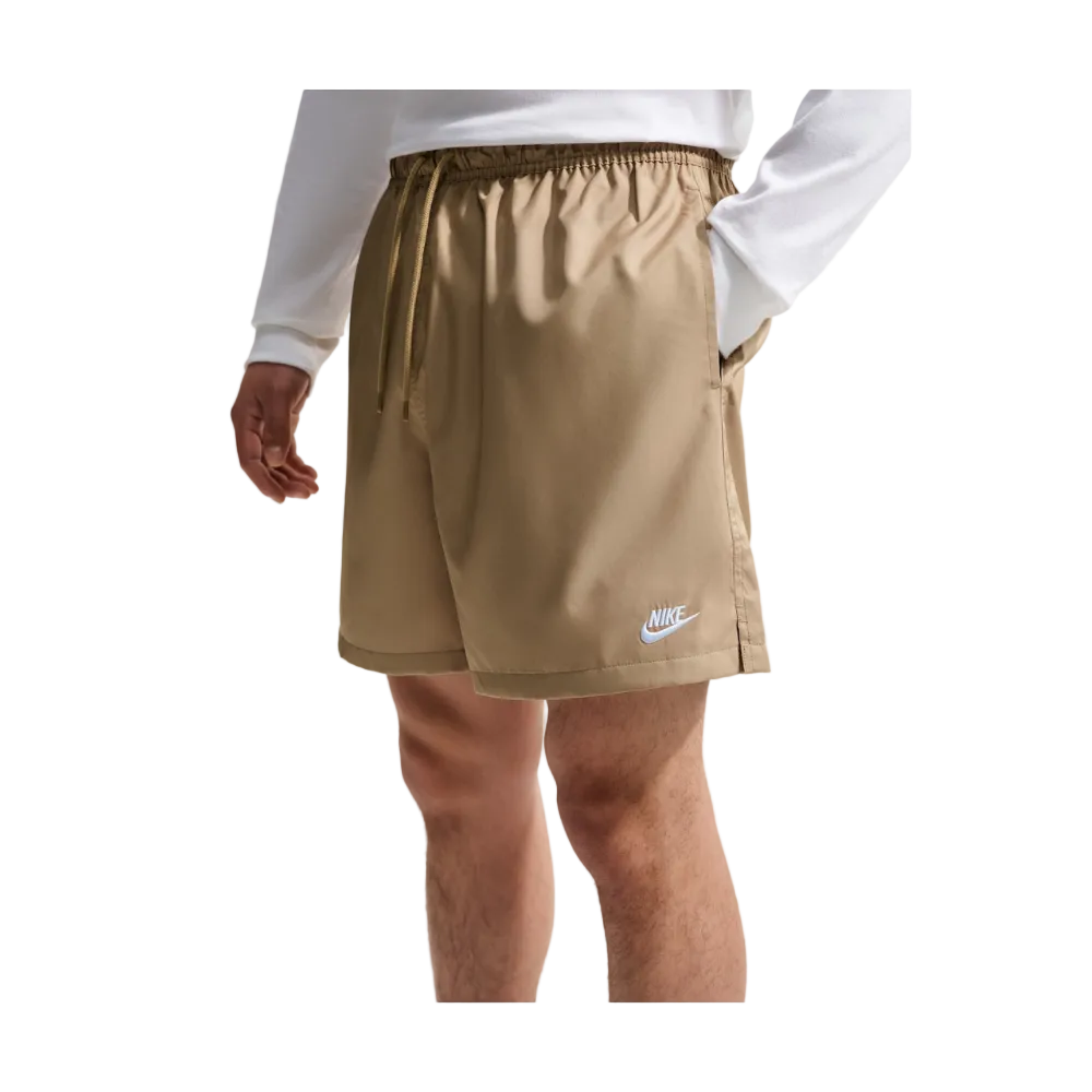A person wearing Nike athletic shorts in a warm khaki-tan color, paired with a white long-sleeve top. The photo is cropped from the chest down, showing the shorts, hands, and legs against a light gray background. 