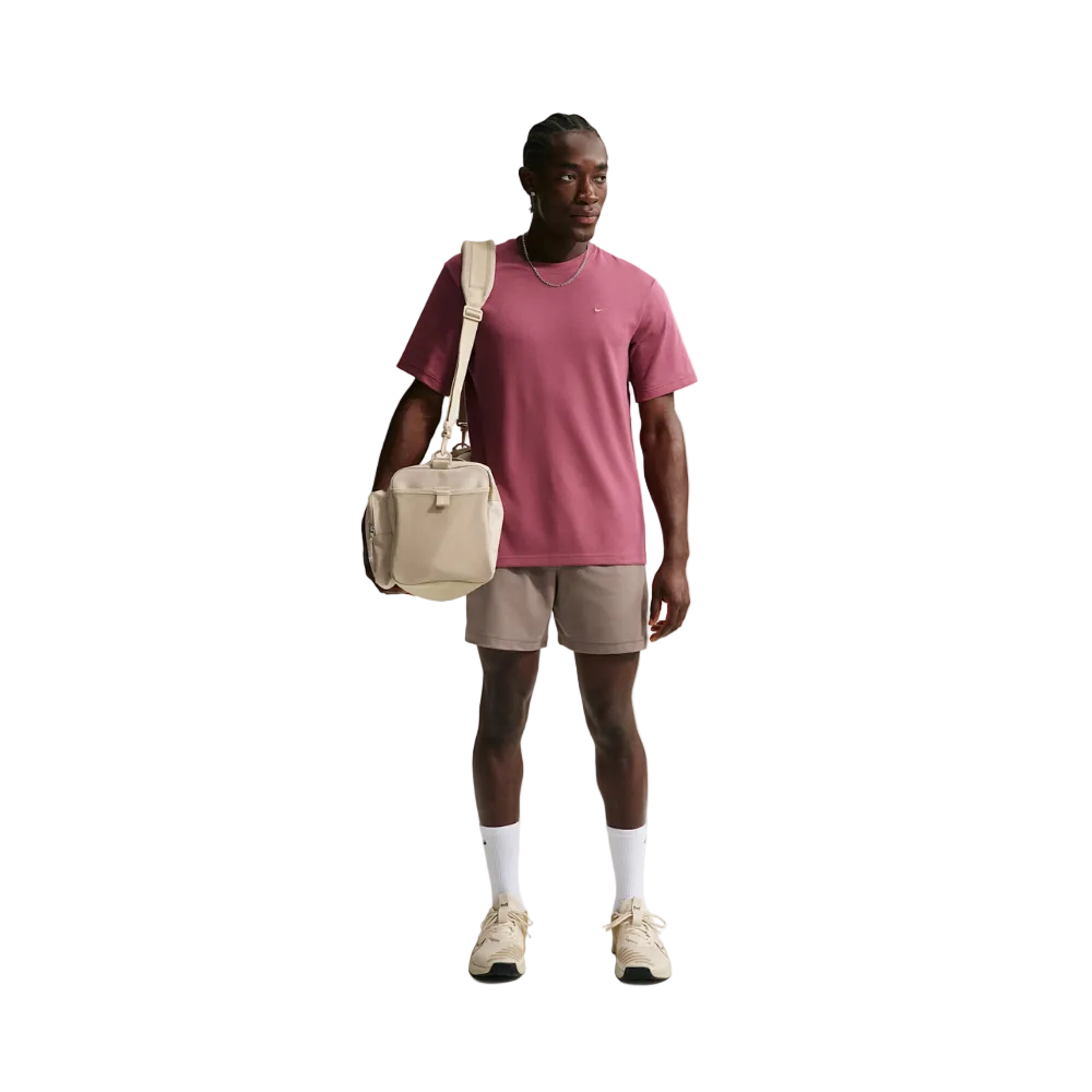 A male model stands against a light gray studio background wearing a dusty rose t-shirt, beige shorts, white socks, and beige sneakers, while carrying a beige shoulder bag.