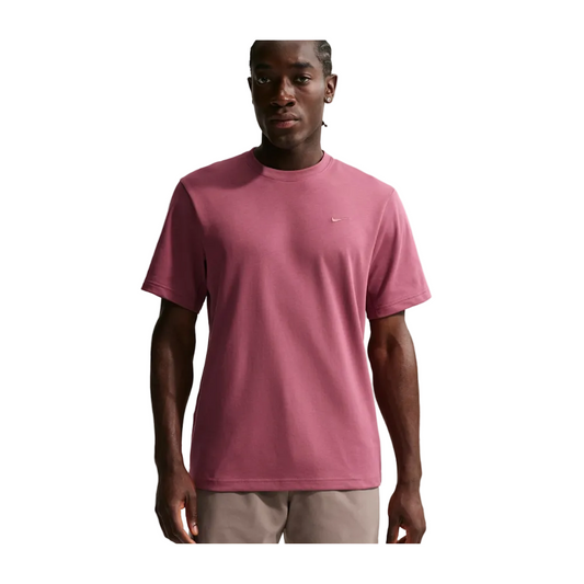 A man wearing a dusty rose pink short-sleeve crewneck t-shirt with a small white Nike swoosh on the left chest, paired with beige pants, set against a solid light gray background.
