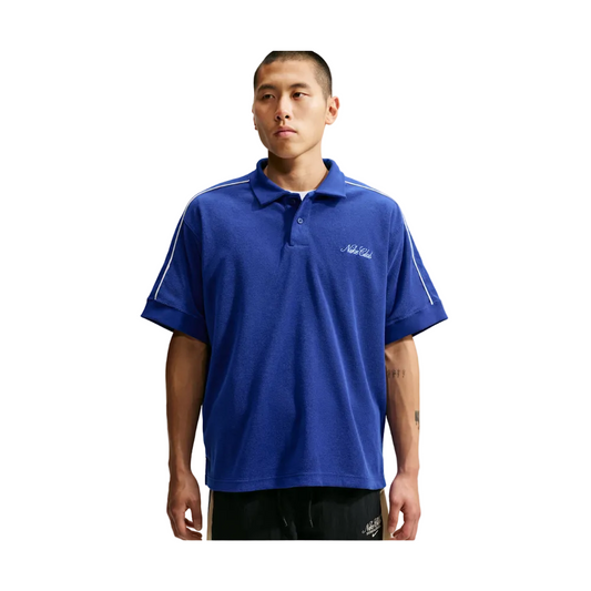 A man wearing a royal blue short-sleeve polo shirt with white stripe details on the shoulders and a small white script logo on the left chest, set against a light gray background.