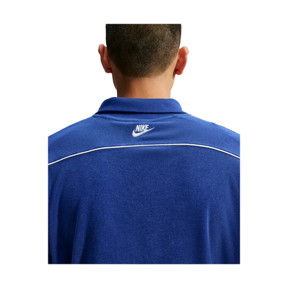 Back view of a person wearing a blue Nike polo shirt with a white logo on the collar and a thin white stripe across the shoulders, set against a light gray background.
