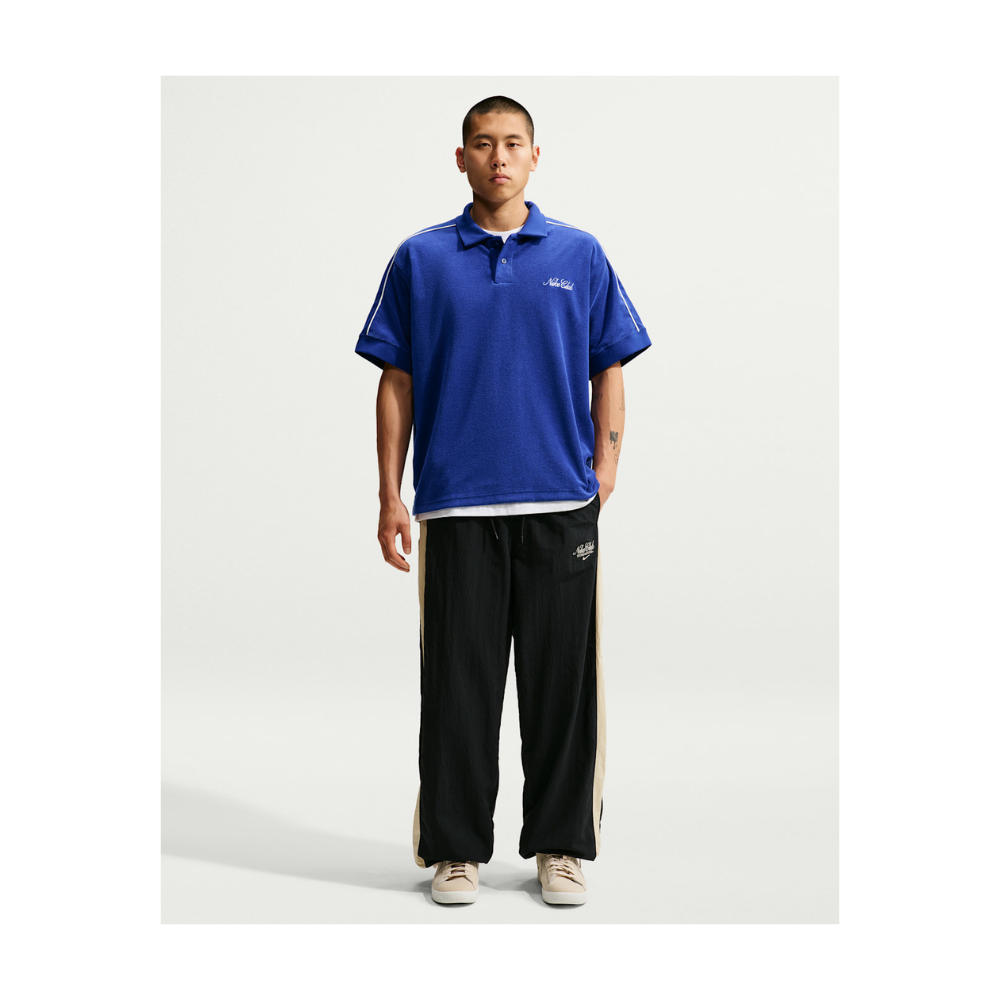 A male model stands against a light gray studio background wearing a royal blue short-sleeve polo shirt with white trim and a small chest logo, paired with black track pants featuring beige side stripes and white sneakers.