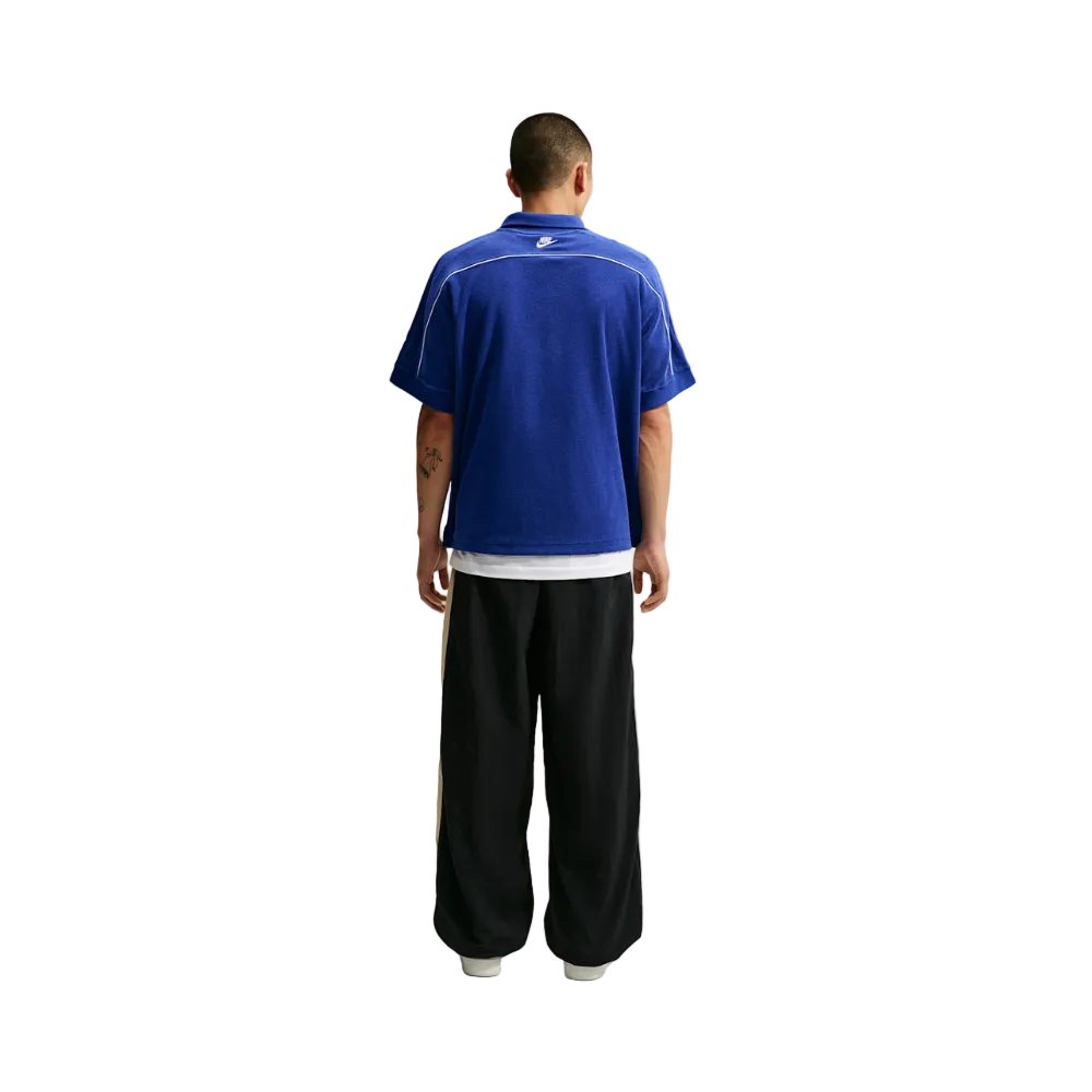 A man viewed from behind wearing a royal blue short-sleeve collared shirt with white piping and black wide-leg pants, standing against a light gray studio background.