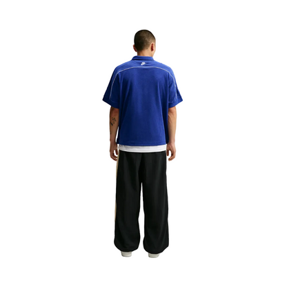 A man viewed from behind wearing a royal blue short-sleeve collared shirt with white piping and black wide-leg pants, standing against a light gray studio background.