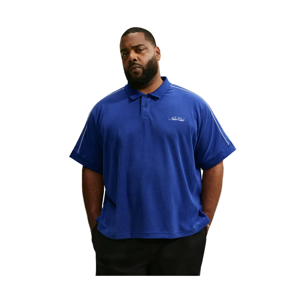 A man wearing a royal blue short-sleeve polo shirt with white piping details on the shoulders and a small white script logo on the left chest, paired with black pants, set against a light gray background.