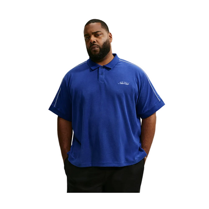 A man wearing a royal blue short-sleeve polo shirt with white piping details on the shoulders and a small white script logo on the left chest, paired with black pants, set against a light gray background.