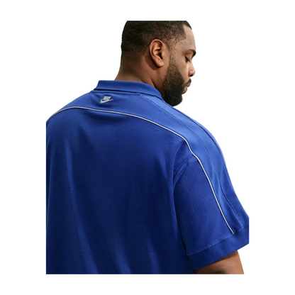 A man wearing a royal blue short-sleeve polo shirt with white piping and a small white Nike logo on the collar, shown from a side-back angle against a light gray background.