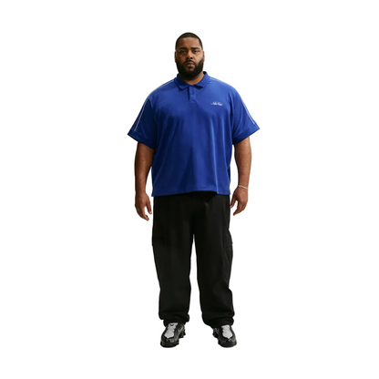 A man modeling a royal blue short-sleeve polo shirt with a small white embroidered logo on the left chest, paired with black pants and black-and-white sneakers, against a light gray studio background.