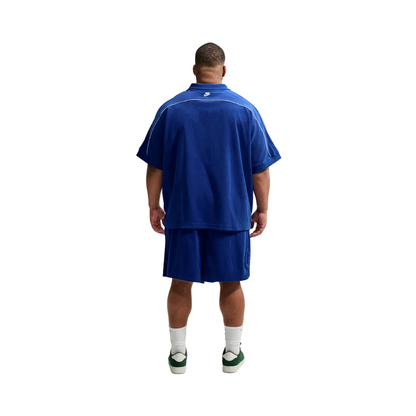 A man viewed from behind wearing a royal blue short-sleeve polo shirt and matching shorts, with white socks and green sneakers, against a plain light gray studio background.