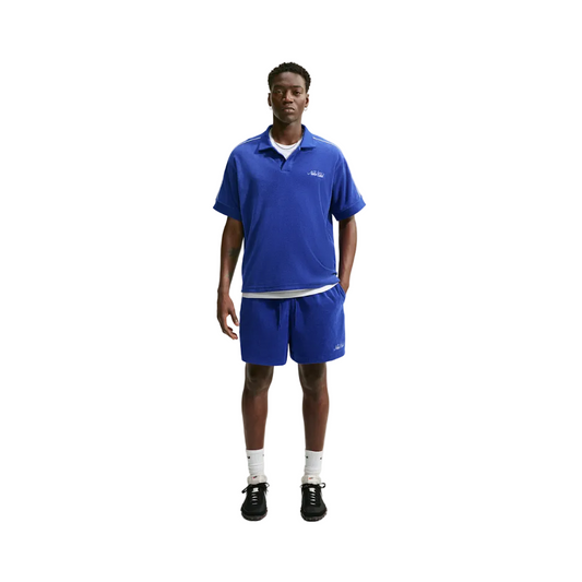 A man modeling a royal blue short-sleeve polo shirt and matching shorts set, styled with white crew socks and black sneakers against a light gray studio background.