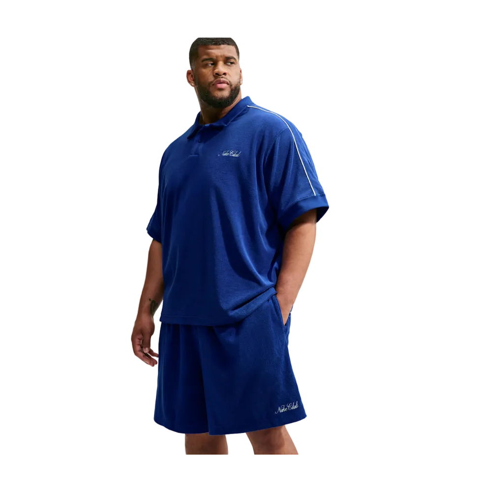 A man modeling a matching royal blue athletic set consisting of a short-sleeve polo shirt and shorts, featuring white embroidered logos on the chest and hem, set against a light gray background.