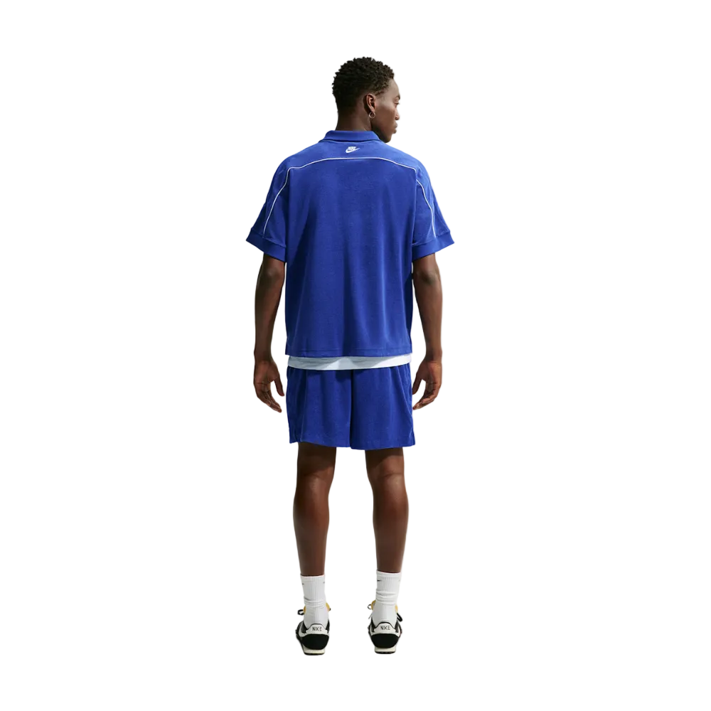 A man viewed from the back wearing a royal blue short-sleeve athletic polo shirt and matching shorts, with white socks and black-and-white sneakers, against a light gray studio background.