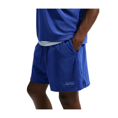 A person wearing a matching royal blue lounge set consisting of a short-sleeve t-shirt and drawstring shorts with a white undershirt peeking out at the hem. The shorts feature a small white embroidered script logo on the lower left leg.