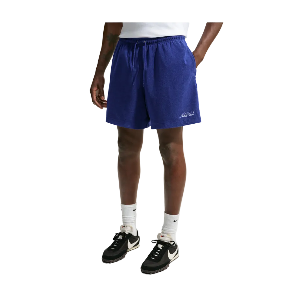 A person wearing royal blue drawstring shorts with a small white script logo near the lower left hem, paired with white socks and black-and-white sneakers, photographed against a light gray studio background.