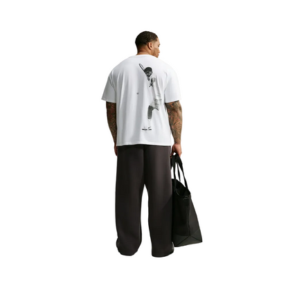 A man stands with his back to the camera wearing a white oversized T-shirt with a large black graphic print on the back, loose dark pants, and holding a black tote bag against a plain light gray studio background.