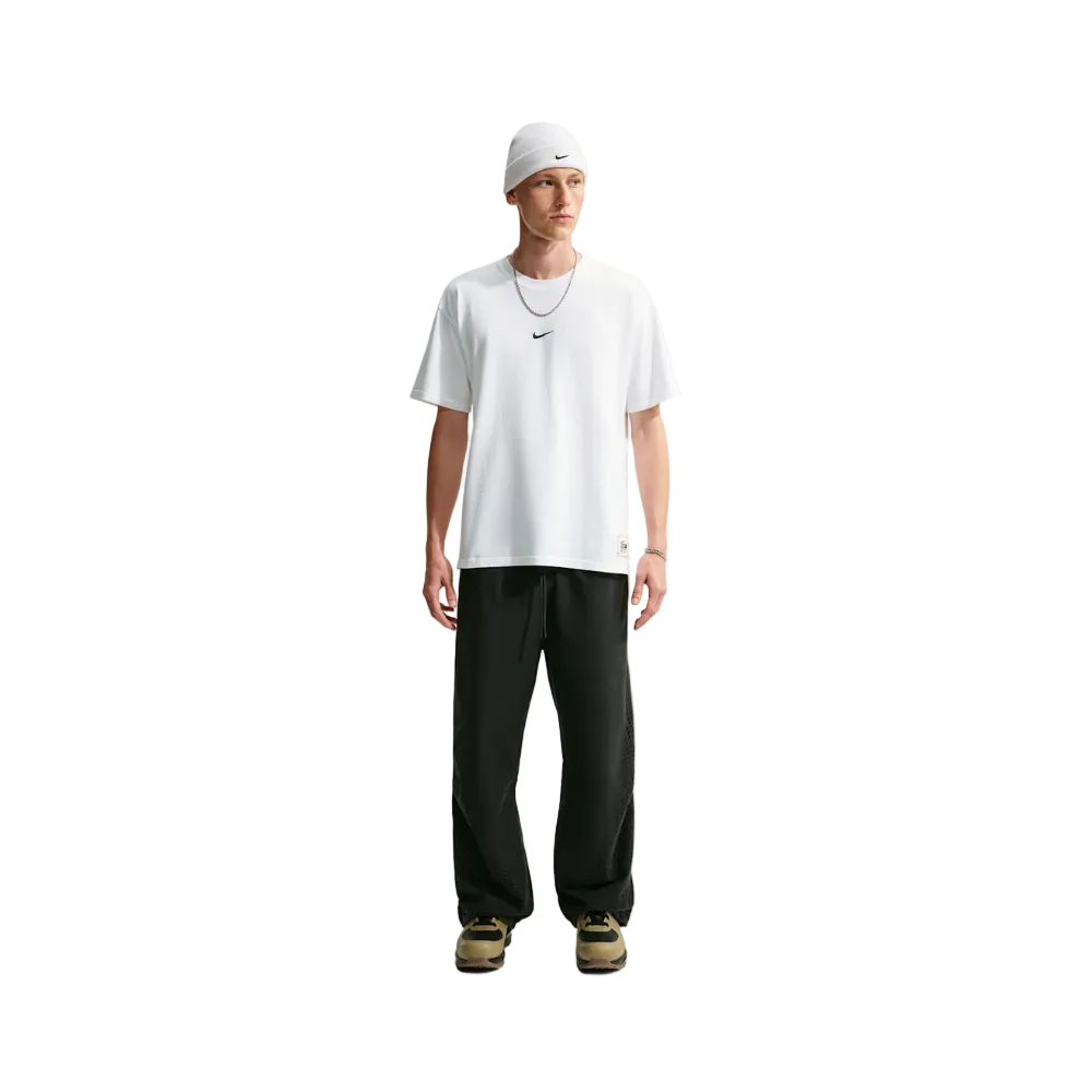 A male model stands against a light gray studio background wearing a white Nike t-shirt, black sweatpants, a white beanie, and sneakers.
