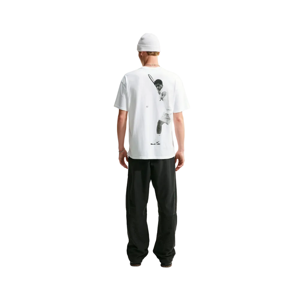 A man viewed from behind wearing a white t-shirt with a large black tennis player graphic on the back, black sweatpants, and a white beanie, standing against a light gray studio background.