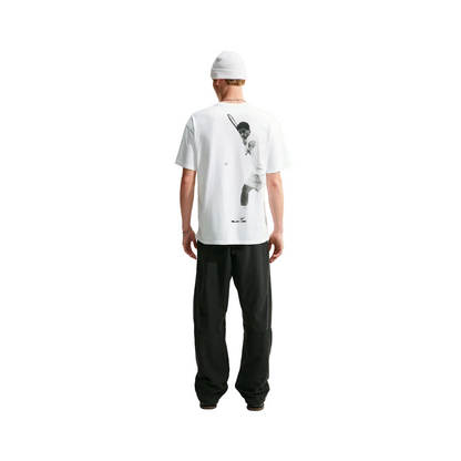 A man viewed from behind wearing a white t-shirt with a large black tennis player graphic on the back, black sweatpants, and a white beanie, standing against a light gray studio background.