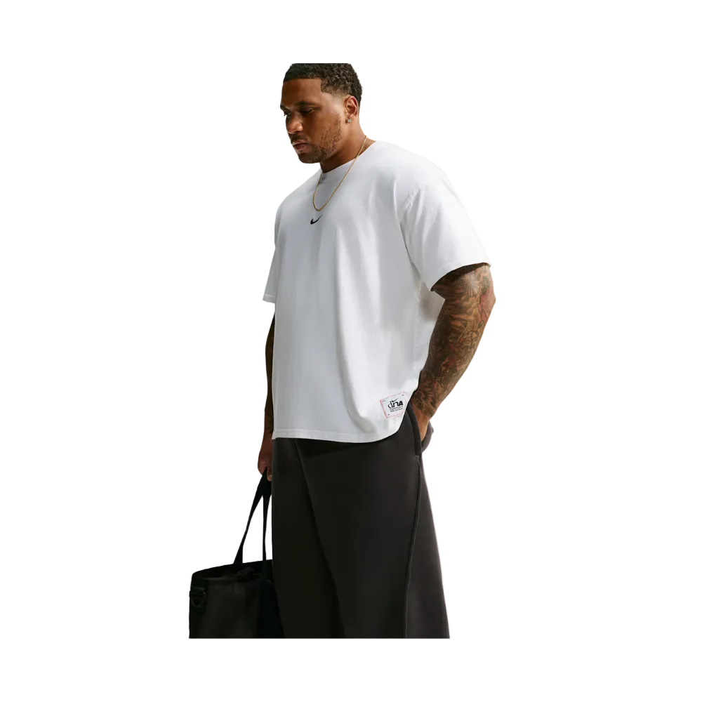 A man wearing a white short-sleeve t-shirt with a small black Nike logo on the chest, paired with black pants, holding a black tote bag against a light gray background.
