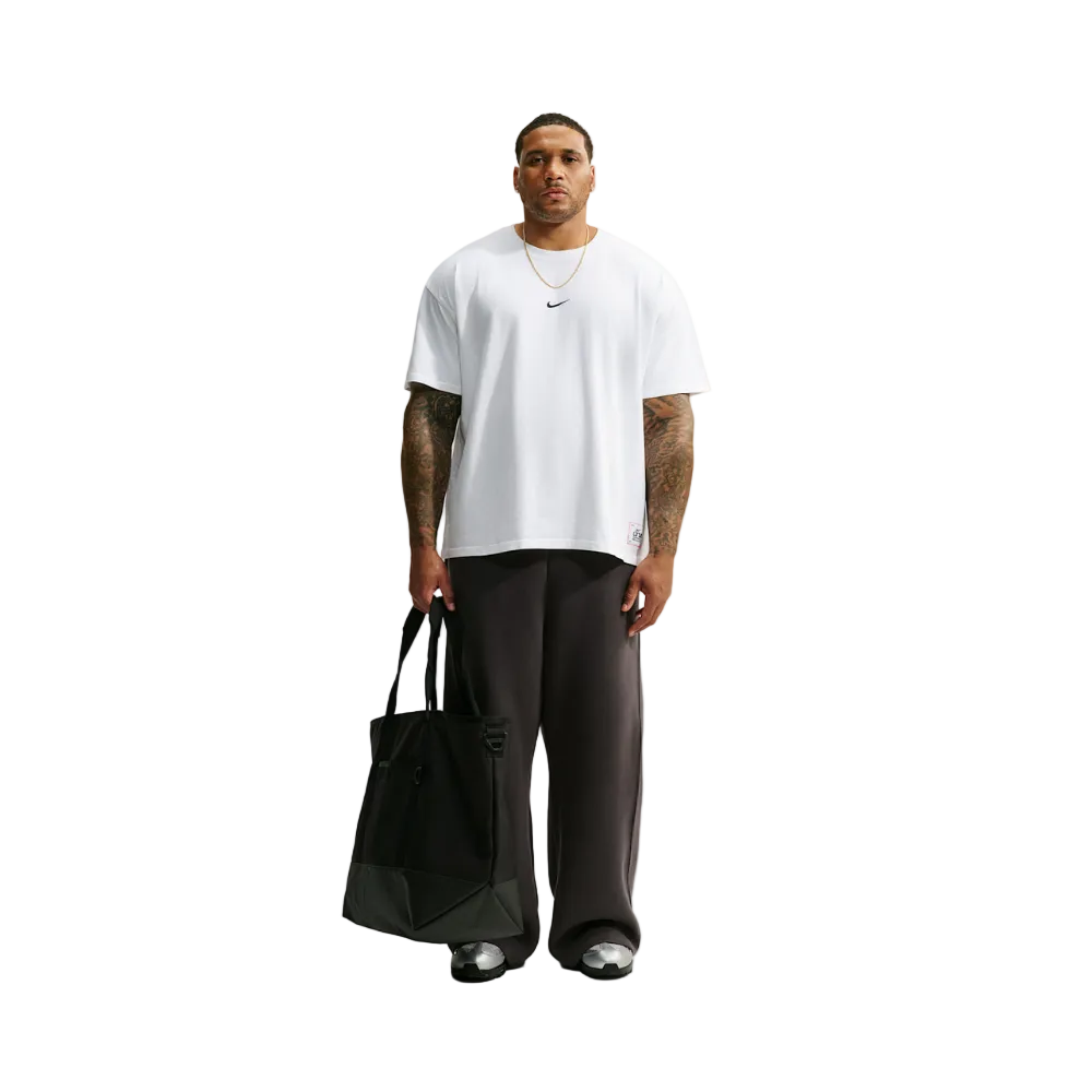 A man stands against a light gray studio background wearing an oversized white Nike T-shirt, loose dark charcoal pants, and black sneakers, while holding a large black tote bag.