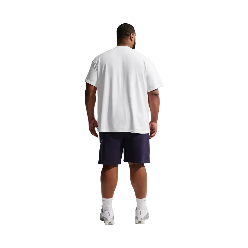 A man viewed from behind wearing an oversized white t-shirt, dark navy athletic shorts, white socks, and white sneakers, standing against a light gray studio background.