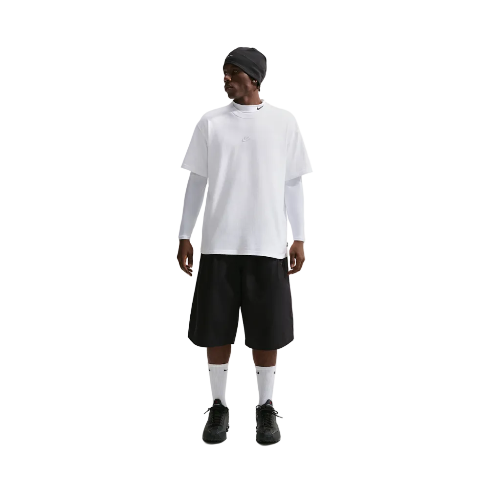 A model stands against a light gray studio background wearing an oversized white short-sleeve t-shirt layered over a white long-sleeve shirt, black shorts, white socks, and black sneakers, with a black beanie on.