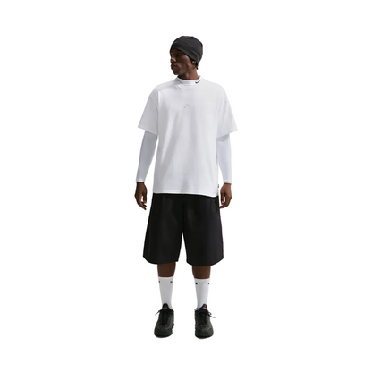 A model stands against a light gray studio background wearing an oversized white short-sleeve t-shirt layered over a white long-sleeve shirt, black shorts, white socks, and black sneakers, with a black beanie on.