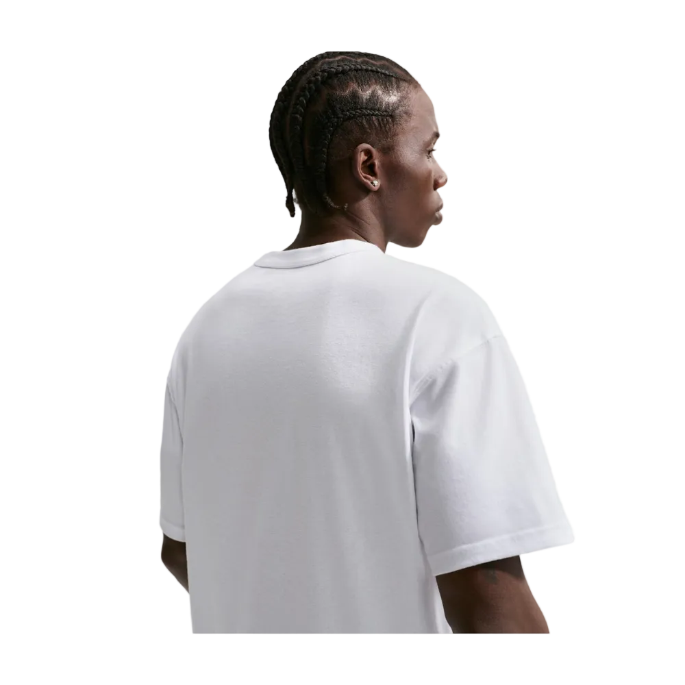 A man with braided hair is shown from the back wearing an oversized white short-sleeve t-shirt, photographed against a light gray studio background.